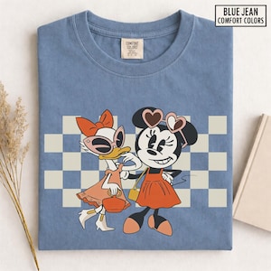 Pu&ograve; includere: T-shirt blu Comfort Colors con grafica retr&ograve; di Minnie e Paperina. Minnie indossa occhiali a forma di cuore e un vestito arancione, mentre Paperina ha occhiali da sole e un abito abbinato. Lo sfondo presenta un motivo a scacchi.