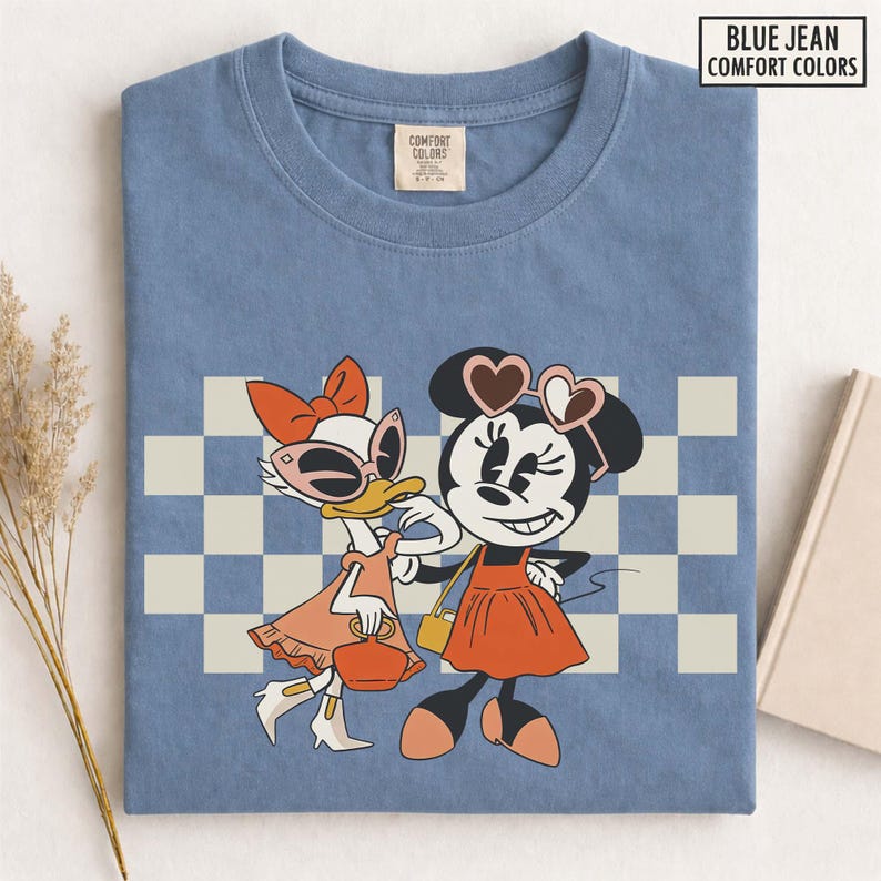 Pu&ograve; includere: T-shirt blu Comfort Colors con grafica retr&ograve; di Minnie e Paperina. Minnie indossa occhiali a forma di cuore e un vestito arancione, mentre Paperina ha occhiali da sole e un abito abbinato. Lo sfondo presenta un motivo a scacchi.
