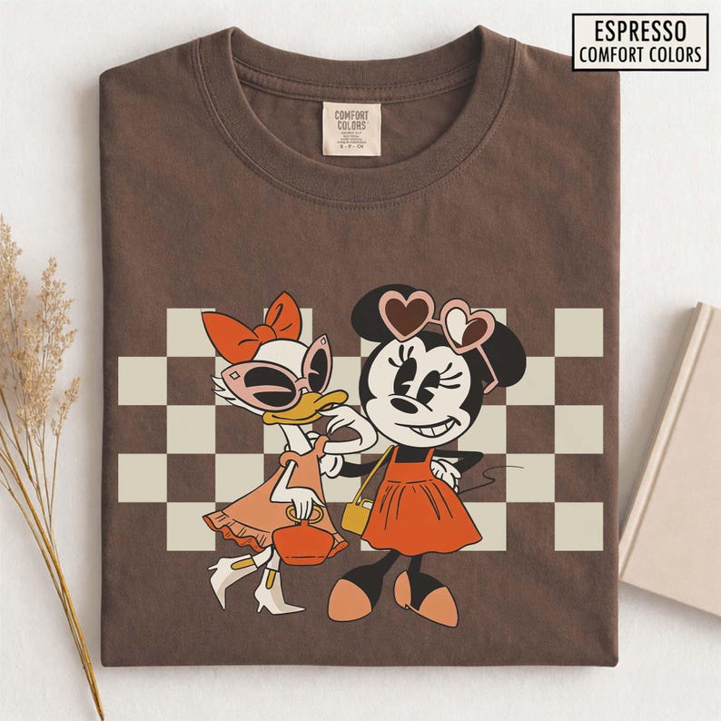 Pu&ograve; includere: T-shirt Comfort Colors color espresso con un'illustrazione di Minnie Mouse e Paperina. Minnie indossa occhiali da sole a forma di cuore e un vestito arancione. Paperina indossa occhiali da sole e un vestito arancione. Lo sfondo ha un motivo a scacchi.