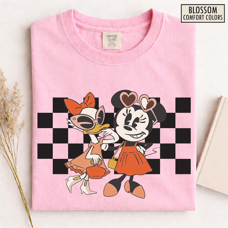 Pu&ograve; includere: T-shirt rosa chiaro con una grafica di Minnie Mouse e Paperina in abiti alla moda e occhiali da sole, su uno sfondo a scacchi bianco e nero. La maglietta ha il testo "Blossom Comfort Colors".