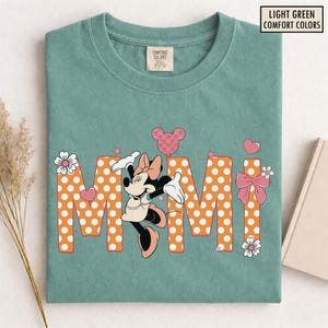 May include: Light green t-shirt with the word "MIMI" in orange polka dot letters, featuring a Minnie Mouse graphic. The design includes hearts, stars, and bows. The shirt has a crew neck and a Comfort Colors tag.