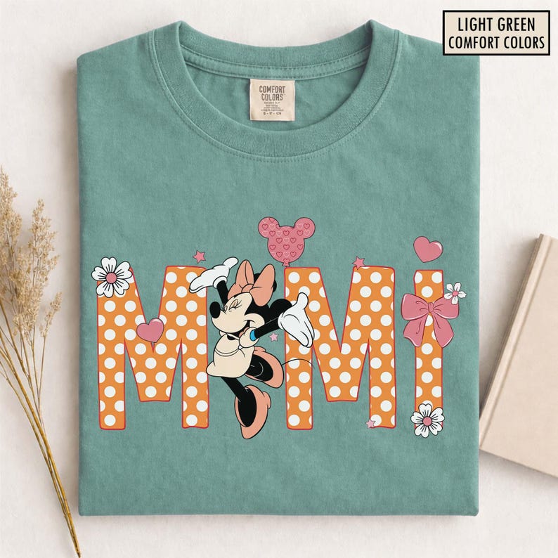 May include: Light green t-shirt with the word "MIMI" in orange polka dot letters, featuring a Minnie Mouse graphic. The design includes hearts, stars, and bows. The shirt has a crew neck and a Comfort Colors tag.