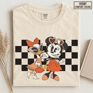 Pu&ograve; includere: T-shirt Ivory Comfort Colors con un'illustrazione di Minnie Mouse e Paperina. Minnie indossa occhiali da sole a forma di cuore e un vestito arancione. Paperina indossa occhiali da sole e un vestito arancione. Un motivo a scacchi bianchi e neri &egrave; dietro i personaggi.