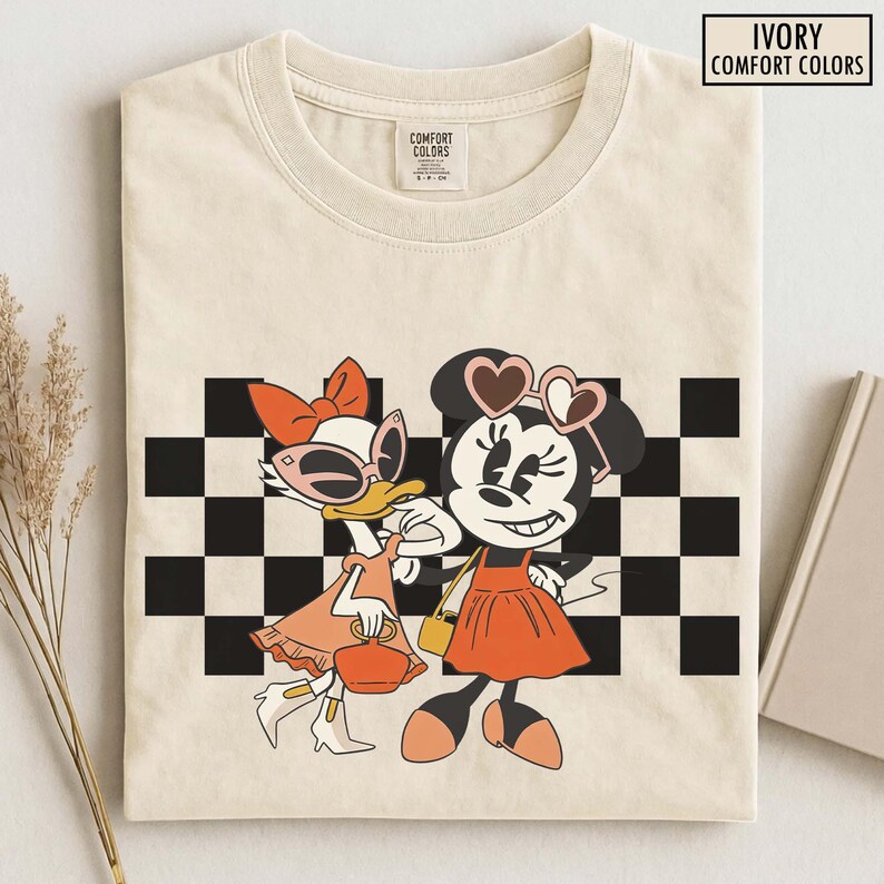 Pu&ograve; includere: T-shirt Ivory Comfort Colors con un'illustrazione di Minnie Mouse e Paperina. Minnie indossa occhiali da sole a forma di cuore e un vestito arancione. Paperina indossa occhiali da sole e un vestito arancione. Un motivo a scacchi bianchi e neri &egrave; dietro i personaggi.