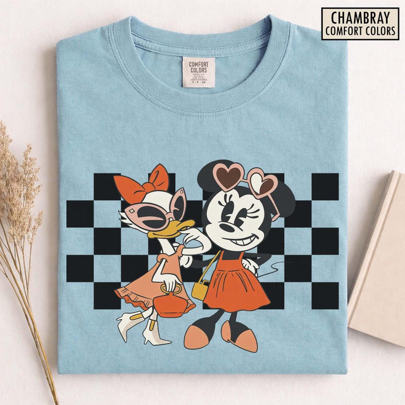 Pu&ograve; includere: T-shirt azzurra con grafica di Paperina e Minnie. Paperina indossa occhiali da sole e un vestito arancione, mentre Minnie indossa occhiali da sole a forma di cuore e un vestito arancione. Lo sfondo presenta un motivo a scacchi bianchi e neri. Il testo "CHAMBRAY COMFORT COLORS" &egrave; in alto a destra.
