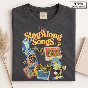 May include: A charcoal gray t-shirt featuring the text "Sing Along Songs" in yellow and orange, with Disney characters and cassette tapes. The shirt includes the text "Disneyland Fun" and "It's a Small World". The brand is "Pepper Comfort Colors".