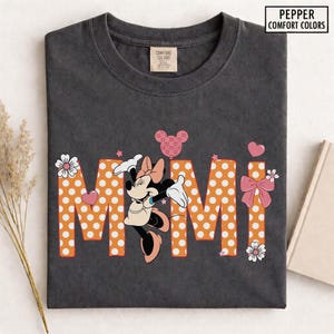 May include: Dark gray t-shirt featuring the word "Mimi" in orange polka-dot letters, with a Minnie Mouse graphic. The shirt includes a pink bow, hearts, and flowers. The text "Pepper Comfort Colors" is in the upper right corner.