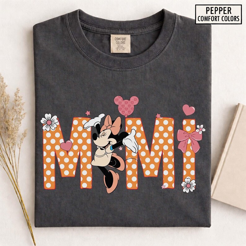 May include: Dark gray t-shirt featuring the word "Mimi" in orange polka-dot letters, with a Minnie Mouse graphic. The shirt includes a pink bow, hearts, and flowers. The text "Pepper Comfort Colors" is in the upper right corner.