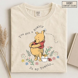 May include: Ivory Comfort Colors t-shirt featuring a Winnie the Pooh graphic. The illustration shows Pooh with his arms crossed, a honey pot, and the text "I've got a Rumbly in my Tumbly..."