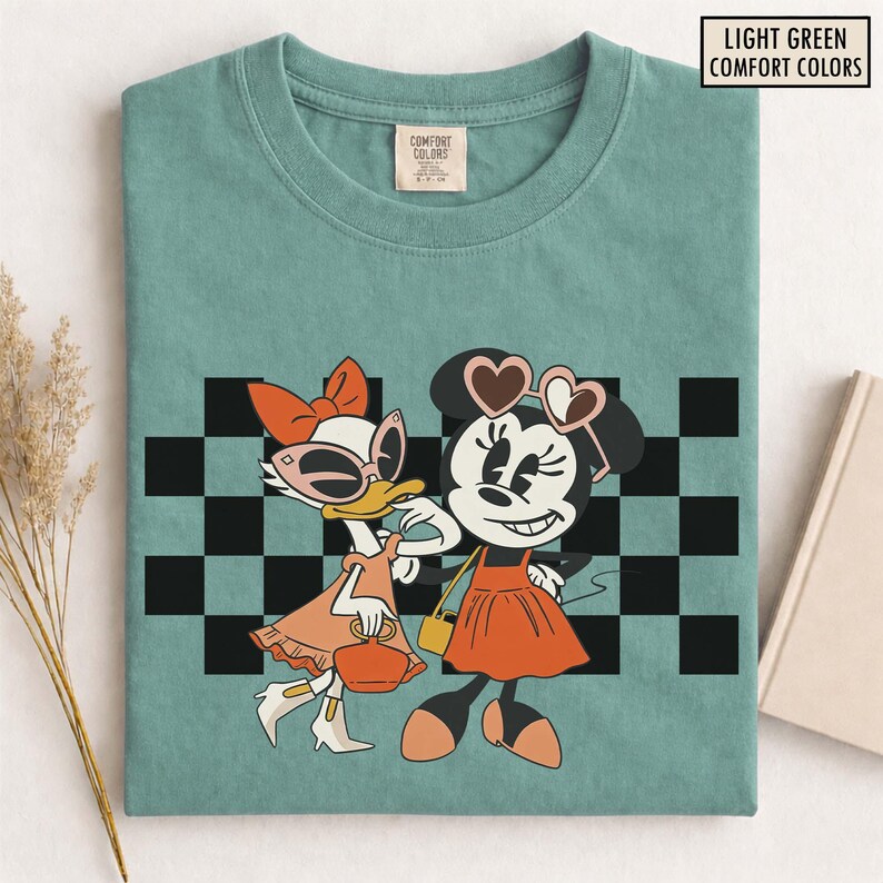 Pu&ograve; includere: T-shirt verde chiaro con una grafica di Minnie Mouse e Paperina, entrambe con occhiali da sole e abiti alla moda, su uno sfondo a scacchi in bianco e nero. Il testo "LIGHT GREEN COMFORT COLORS" si trova nell'angolo in alto a destra.