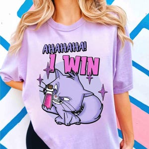Puede incluir: Camiseta lila claro con un gato de dibujos animados sosteniendo una botella de poción rosa. El texto "AHAHAHA! I WIN" está encima del gato en morado y rosa. El diseño incluye estrellas.