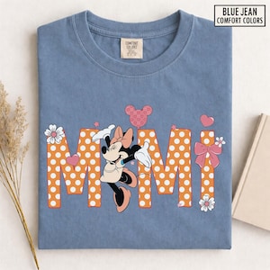 May include: A blue jean Comfort Colors t-shirt featuring Minnie Mouse and the word "Mimi" in orange polka-dot letters. The design includes hearts, stars, and bows. The shirt has a crew neck and a small tag on the upper chest.