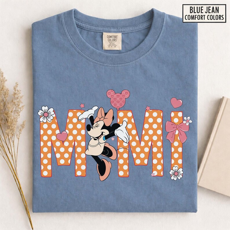 May include: A blue jean Comfort Colors t-shirt featuring Minnie Mouse and the word "Mimi" in orange polka-dot letters. The design includes hearts, stars, and bows. The shirt has a crew neck and a small tag on the upper chest.