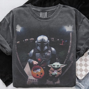 Pode incluir: Uma t-shirt de gola redonda cinzento-carvão com uma impressão gráfica de uma personagem mandaloriana com capacete a segurar um balde de doces de abóbora laranja, ao lado de uma pequena criatura verde com uma túnica castanha.