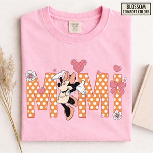 May include: Pink t-shirt with the word "MIMI" in orange polka dot letters. Minnie Mouse is featured in the center, with a pink bow, heart-shaped balloon, and floral accents. The shirt has the text "BLOSSOM COMFORT COLORS".