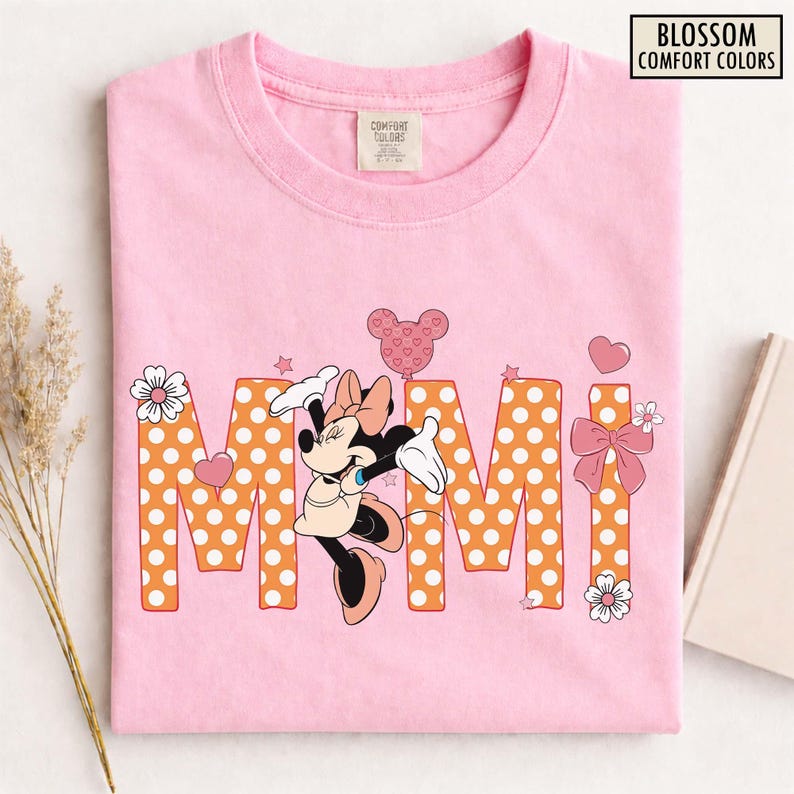 May include: Pink t-shirt with the word "MIMI" in orange polka dot letters. Minnie Mouse is featured in the center, with a pink bow, heart-shaped balloon, and floral accents. The shirt has the text "BLOSSOM COMFORT COLORS".