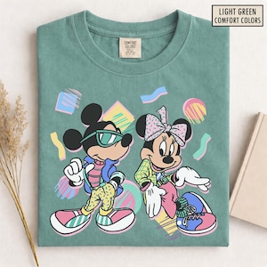 Puede incluir: Camiseta verde claro Comfort Colors con Mickey y Minnie Mouse con atuendos de los 80. Mickey lleva gafas de sol, una chaqueta azul y pantalones amarillos. Minnie tiene un lazo y una chaqueta a cuadros verde. Formas geométricas coloridas.