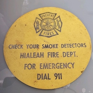 May include: A circular yellow sticker featuring a black fire department emblem at the top with the text PREVENT FIRES. Below, the text reads CHECK YOUR SMOKE DETECTORS, HIALEAH FIRE DEPT., FOR EMERGENCY DIAL 911 in a bold, black, sans-serif font.