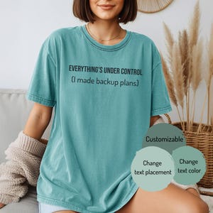May include: A teal t-shirt featuring the text "EVERYTHING'S UNDER CONTROL (I made backup plans)" in black. The shirt has a crew neck and short sleeves. The image also includes the words "Customizable", "Change text placement", and "Change text color."