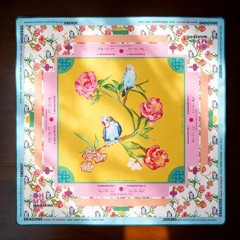 Peut inclure: Un tapis de Mahjong carr&eacute; avec un centre jaune, deux perruches bleues perch&eacute;es sur une branche fleurie de pivoines roses. La bordure pr&eacute;sente un motif floral, le texte OH MY MAHJONG et des instructions de jeu comme CHARLESTON 1 et 2.