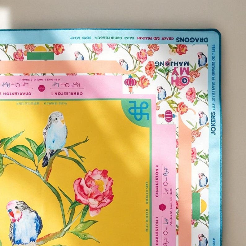 Peut inclure: Un tapis de jeu rectangulaire avec un centre jaune orn&eacute; de perruches bleues et de pivoines roses. La bordure pr&eacute;sente des sections bleu clair, roses et blanches avec des motifs floraux, des lanternes et le texte "OH MY MAHJONG" et "JOICERS".