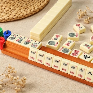 Peut inclure: Un ensemble de tuiles de Mahjong blanc cassé avec des marquages à l'encre rouge, verte et noire, incluant des caractères et des lettres latines, disposées sur un support en bois. Certaines tuiles présentent des motifs floraux et de bambou.