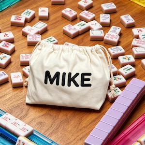 以下が含まれることがあります： 黒い文字でMIKEと刺繍されたクリーム色の巾着袋。その周りには様々なシンボルや色が描かれた麻雀牌が散らばっており、青、ピンク、紫のプラスチック製牌ラックが木製の表面に置かれています。