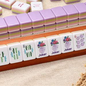 May include: A set of Mahjong tiles featuring a lavender, green, and white layered design. The tiles display various illustrations including bamboo stalks, floral vases labeled Joker, purple flower clusters, and a colorful bird on a branch.