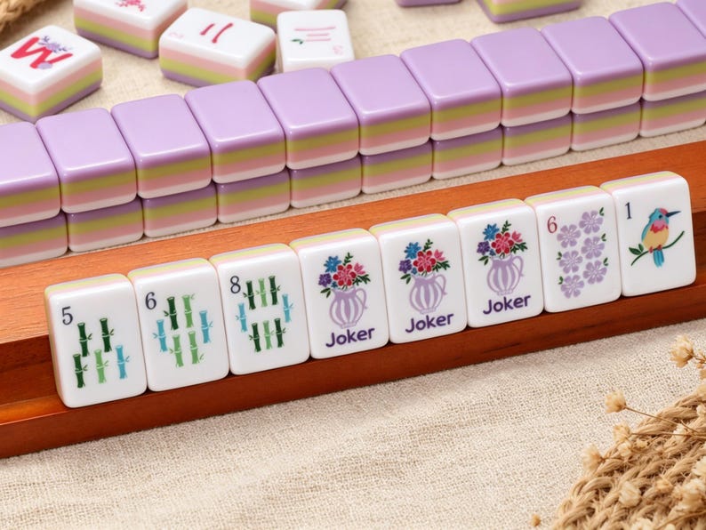 Pu&ograve; includere: Un set di tessere del Mahjong con un design a strati color lavanda, verde e bianco. Le tessere mostrano varie illustrazioni, tra cui steli di bamb&ugrave;, vasi di fiori etichettati Joker, grappoli di fiori viola e un uccellino colorato su un ramo.