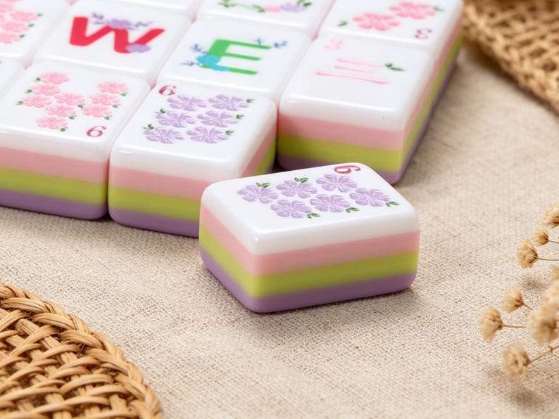 May include: A set of rectangular mahjong tiles featuring a layered design with white, pink, light green, and lavender stripes. The top surfaces display various floral patterns, the letter W, the letter E, and the number 6 in different colors.