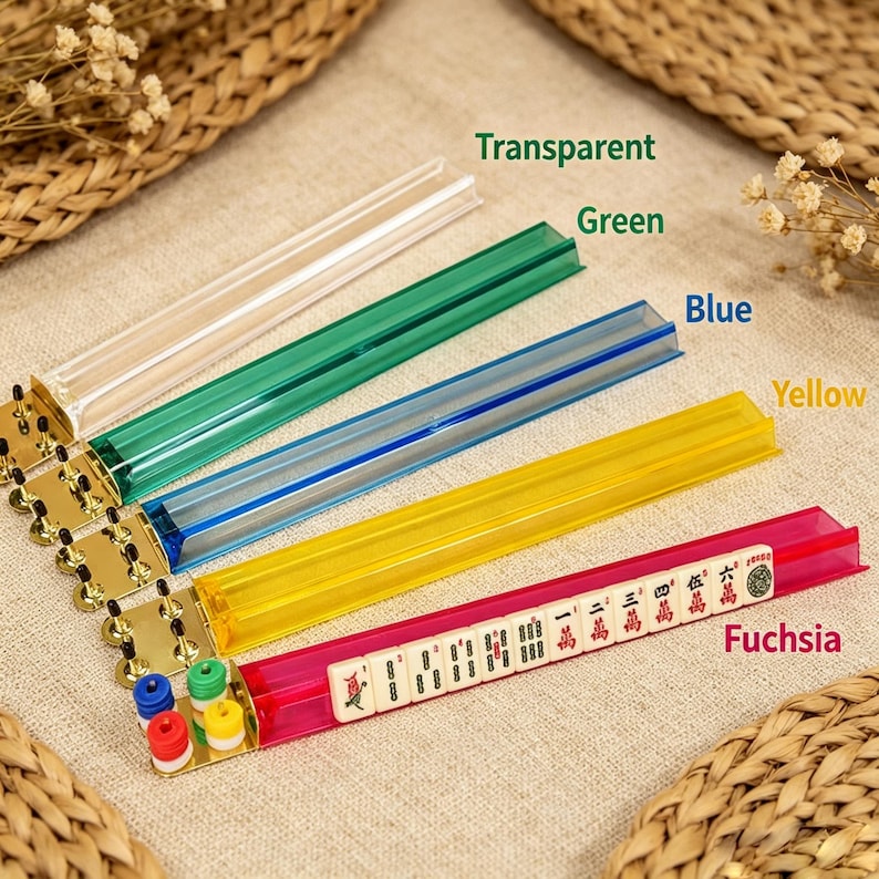 May include: Five long, narrow Mahjong tile racks in transparent, green, blue, yellow, and fuchsia. Each rack features a gold-colored metal end piece with small, colorful cylindrical pegs. The fuchsia rack holds a row of ivory-colored Mahjong tiles.