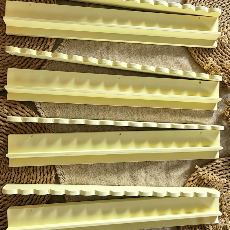 May include: A set of four pale yellow wall-mounted shelves, each featuring a scalloped front edge and a flat back panel with pre-drilled holes for mounting. The shelves have a smooth, matte finish and a narrow ledge for displaying small items.