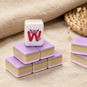 May include: A set of rectangular mahjong tiles featuring a layered color design of purple, light green, and white. One tile stands vertically on top of the others, displaying a red letter W decorated with small purple and blue floral accents.