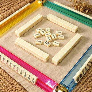 May include: A Mahjong game set featuring four translucent plastic racks in yellow, green, pink, and blue. The set includes numerous off-white tiles, some arranged in rows on the racks and others scattered in the center, displaying various characters and symbols.
