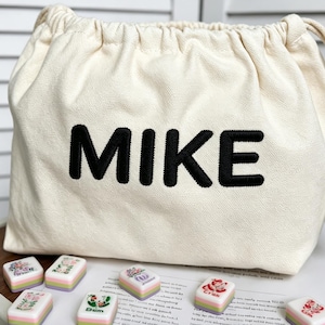 以下が含まれることがあります： 前面に太い黒文字でMIKEと刺繍された、クリーム色のキャンバス地巾着袋。袋の周りには、花柄やグラフィックデザインが施された小さな長方形のタイルがいくつか置かれています。