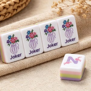 May include: Four rectangular white mahjong tiles featuring a graphic of a purple vase filled with colorful flowers and the word Joker printed in purple. A single white mahjong tile with a large purple letter N decorated with small pink flowers.