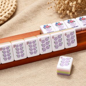 May include: A set of white Mahjong tiles with purple floral patterns and numerical markings, along with four tiles featuring a colorful flower vase graphic and the word Joker, all arranged on a long wooden rack with a grooved surface.