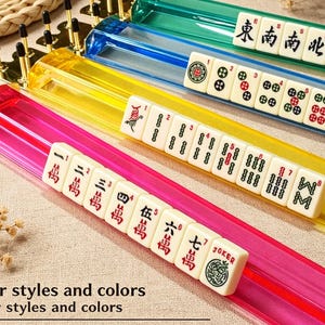 May include: Three translucent Mahjong tile racks in red, yellow, and blue, each featuring gold-toned metal end caps. The racks hold white tiles with various black, red, and green symbols, numbers, and characters. Text reads: Four styles and colors.