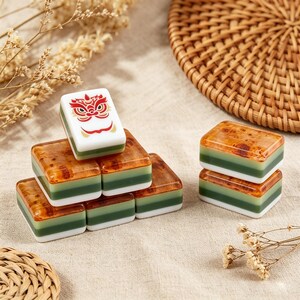 May include: A collection of rectangular mahjong tiles featuring a layered design with a white base, a green middle section, and a glossy amber-colored top. One tile is positioned upright, displaying a red and gold lion dance mask graphic.
