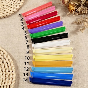 May include: A set of fourteen plastic bag sealing clips arranged in a vertical column, numbered one through fourteen. The clips are available in a variety of solid colors including pink, red, purple, green, black, white, yellow, orange, and blue.