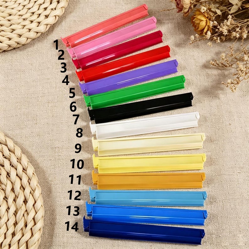 May include: A set of fourteen plastic bag sealing clips arranged in a vertical column, numbered one through fourteen. The clips are available in a variety of solid colors including pink, red, purple, green, black, white, yellow, orange, and blue.