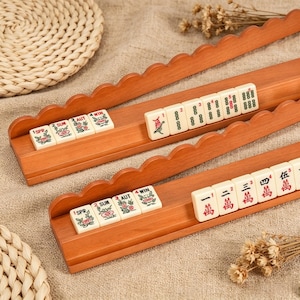 May include: Two wooden Mahjong tile racks with scalloped edges, each holding a row of cream-colored tiles. The top rack displays seasonal flower tiles and numbered tiles. The bottom rack features seasonal tiles and tiles with red Chinese characters.