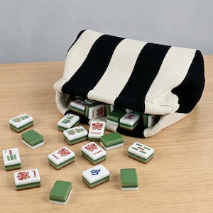May include: A black and cream striped fabric pouch containing a set of rectangular mahjong tiles. The tiles are white with green bases and feature various red, green, and blue symbols, including bamboo patterns, Chinese characters, and dragon motifs.