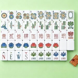 May include: A set of white rectangular mahjong tiles arranged in a grid, featuring colorful illustrations of circles, bamboo, Chinese characters, lotus flowers, compass directions, and joker tiles, with one loose tile placed to the bottom right.