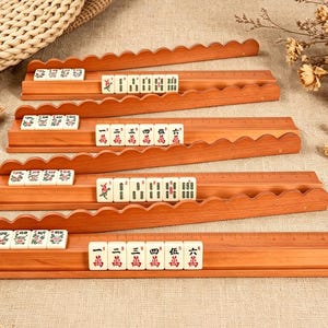 May include: Four wooden mahjong tile racks with scalloped edges, each holding a set of off-white tiles featuring red and green Chinese characters and symbols. The racks are arranged in a stack, displaying a variety of numbered tiles and suits.