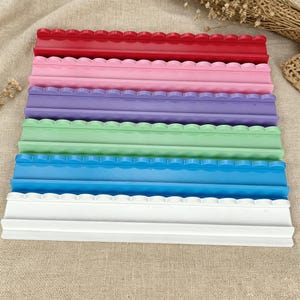 Könnte beinhalten: Ein Set aus sechs rechteckigen Präsentationsständern, die gestapelt sind und jeweils eine gewellte Oberkante aufweisen. Die Farben umfassen Rot, Rosa, Lila, Hellgrün, Blau und Weiß. Jeder Ständer verfügt über eine horizontale Rille.