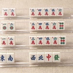 Pode incluir: Quatro suportes de acr&iacute;lico transparente contendo pe&ccedil;as brancas de mahjong. As pe&ccedil;as apresentam v&aacute;rios caracteres chineses, n&uacute;meros e padr&otilde;es circulares verdes em tinta vermelha, azul e verde. Os suportes est&atilde;o dispostos em filas horizontais.