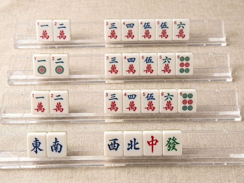 Pode incluir: Quatro suportes de acr&iacute;lico transparente contendo pe&ccedil;as brancas de mahjong. As pe&ccedil;as apresentam v&aacute;rios caracteres chineses, n&uacute;meros e padr&otilde;es circulares verdes em tinta vermelha, azul e verde. Os suportes est&atilde;o dispostos em filas horizontais.