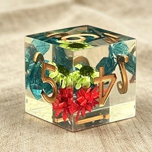 May include: A clear resin cube die featuring embedded dried flowers in shades of red, green, and blue. The die has gold-colored numbers on its faces, including a prominent five and four, with a smooth, polished finish and sharp, defined edges.
