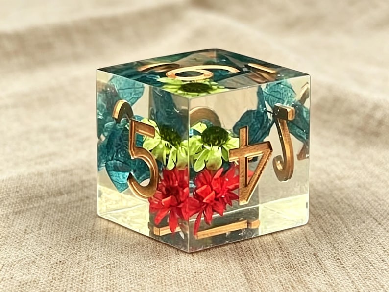 May include: A clear resin cube die featuring embedded dried flowers in shades of red, green, and blue. The die has gold-colored numbers on its faces, including a prominent five and four, with a smooth, polished finish and sharp, defined edges.